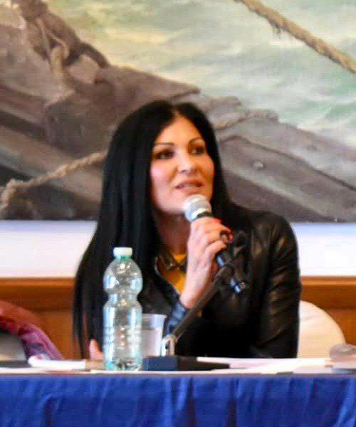 Sonia Buscemi, psicologa clinica, mentre parla a un pubblico durante un evento, con un microfono in mano e una bottiglia d'acqua sulla scrivania.